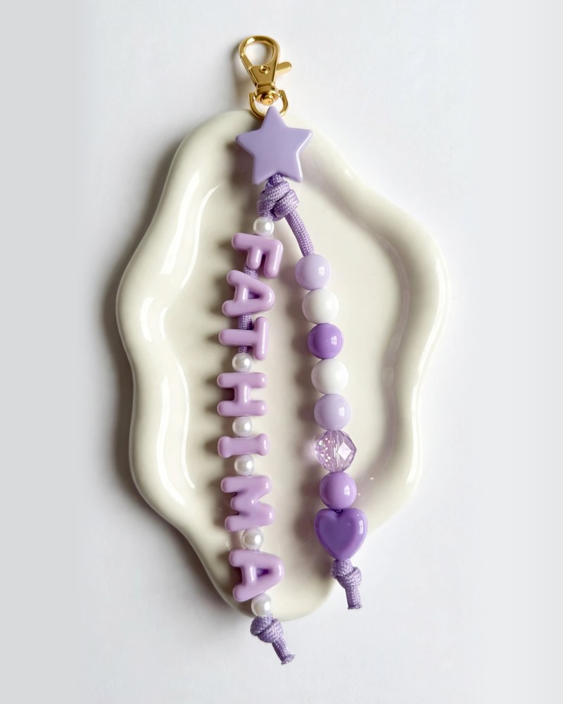 Lavender double-string paracord keychain spelling FATHIMA with pearl beads between each letter, lilac beads, a crystal bead, a purple heart, a lilac star and a gold clasp